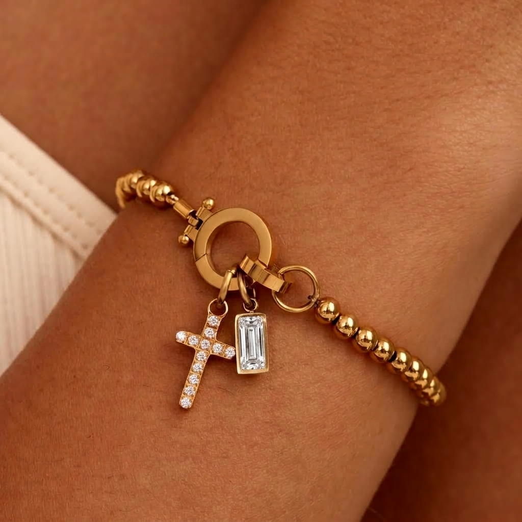 Gold-tone beaded bracelet on a wrist with a sparkling cross charm and a clear baguette-cut stone charm hanging from the round clasp. 21