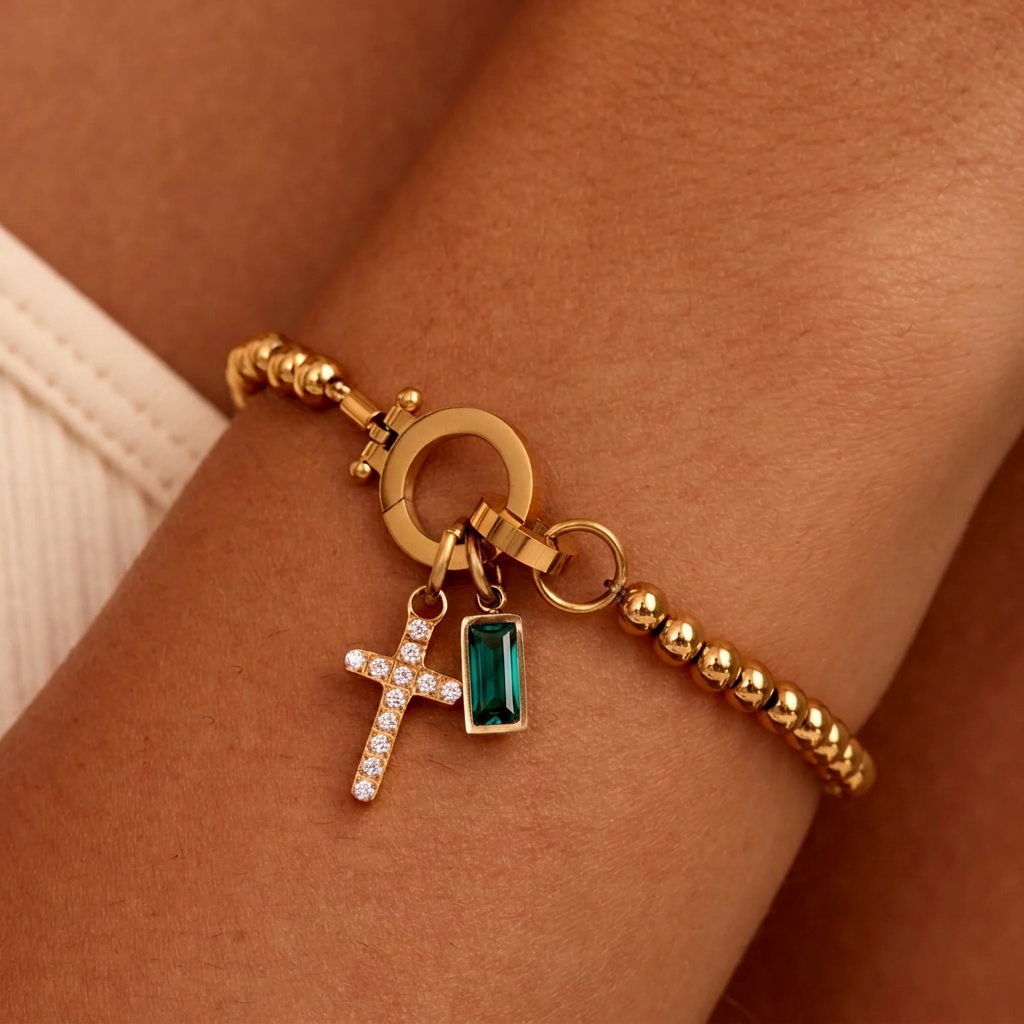 Gold-tone beaded bracelet on a wrist with a sparkling cross charm and a deep green baguette-cut stone charm hanging from the clasp. 23