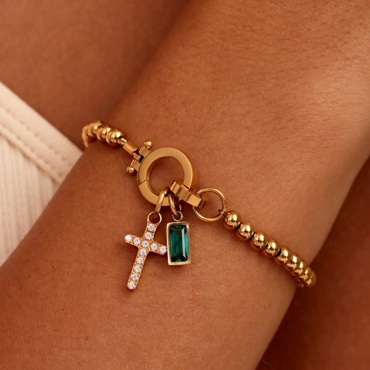 Gold-tone beaded bracelet on a wrist with a sparkling cross charm and a deep green baguette-cut stone charm hanging from the clasp. 23