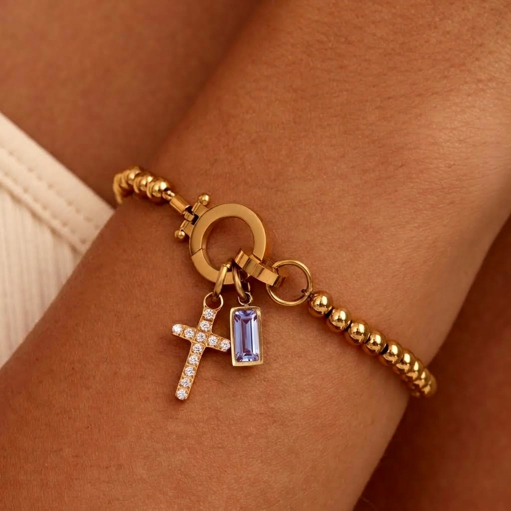 Gold-tone beaded bracelet worn on a wrist, with a sparkling cross charm and a pale lavender baguette-cut stone charm. 25