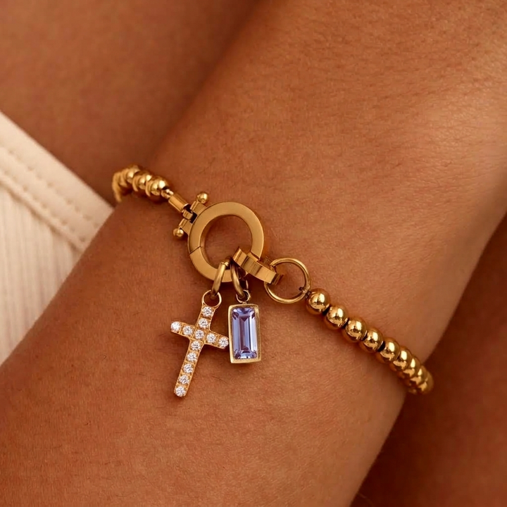 Gold-tone beaded bracelet worn on a wrist, with a sparkling cross charm and a pale lavender baguette-cut stone charm. 25