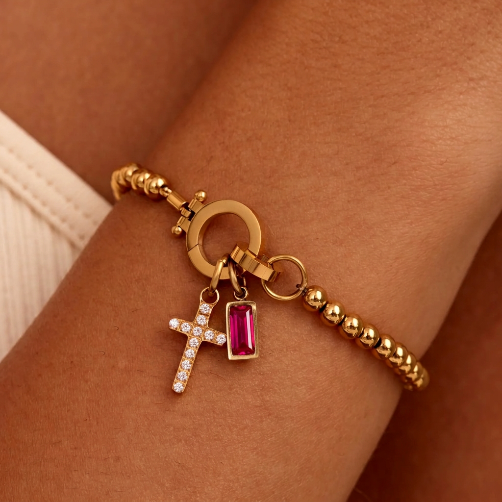 Gold-tone beaded bracelet on a wrist with a sparkling cross charm and a deep pink-red baguette-cut stone charm hanging beside it. 27