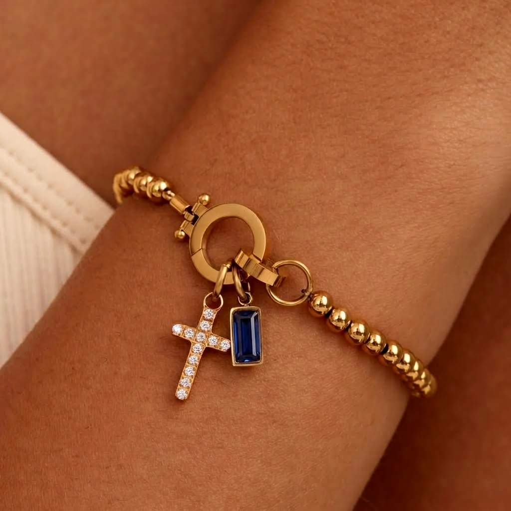 Gold-tone beaded bracelet worn on a wrist, with a sparkling cross charm and a deep blue baguette-cut stone charm. 31