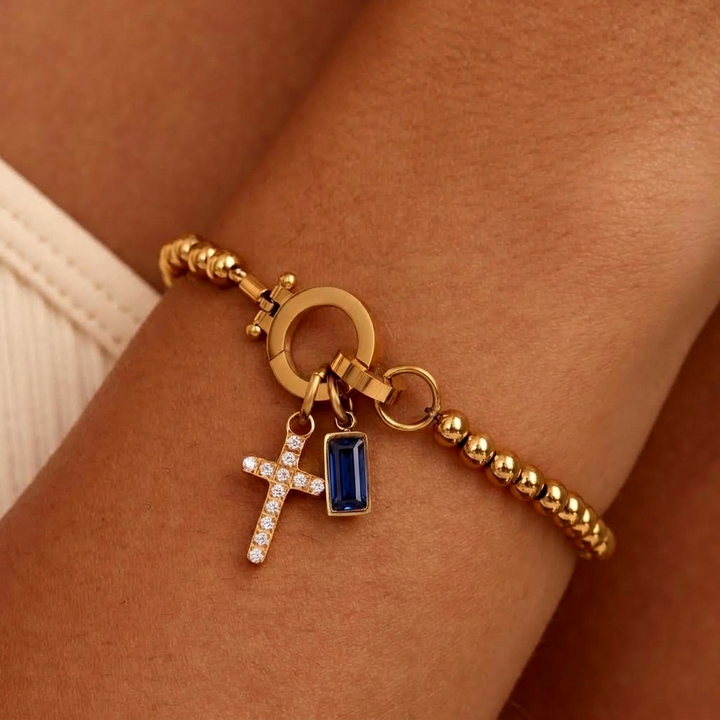 Gold-tone beaded bracelet worn on a wrist, with a sparkling cross charm and a deep blue baguette-cut stone charm. 31