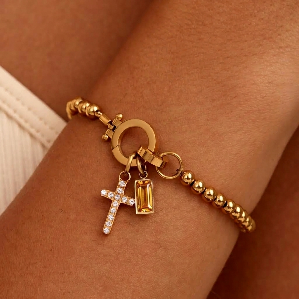 Gold-tone beaded bracelet on a wrist with a sparkling cross charm and a golden amber baguette-cut stone charm at the round clasp. 35