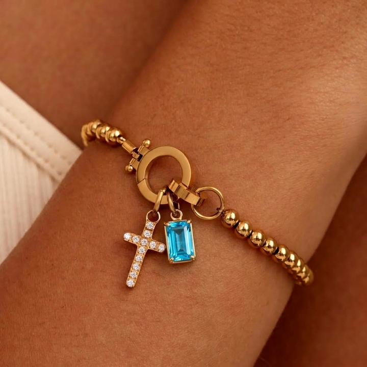Gold-tone beaded bracelet worn on a wrist, with a sparkling cross charm and a larger bright aqua blue stone charm in a prong setting. 37
