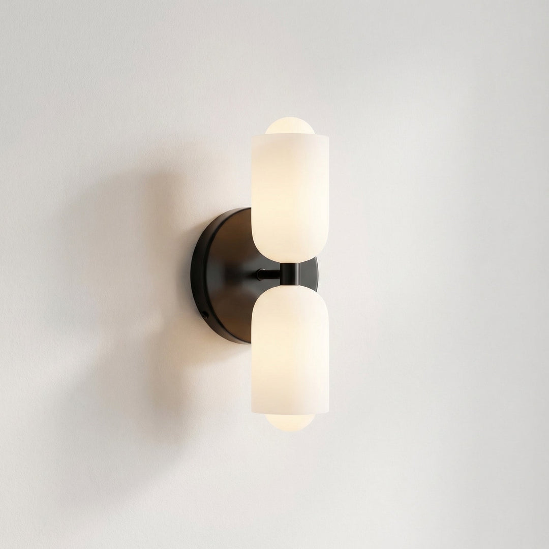 Close-up of the black wall light with two frosted glass shades and a short black arm joining them, glowing on a plain white wall. 8