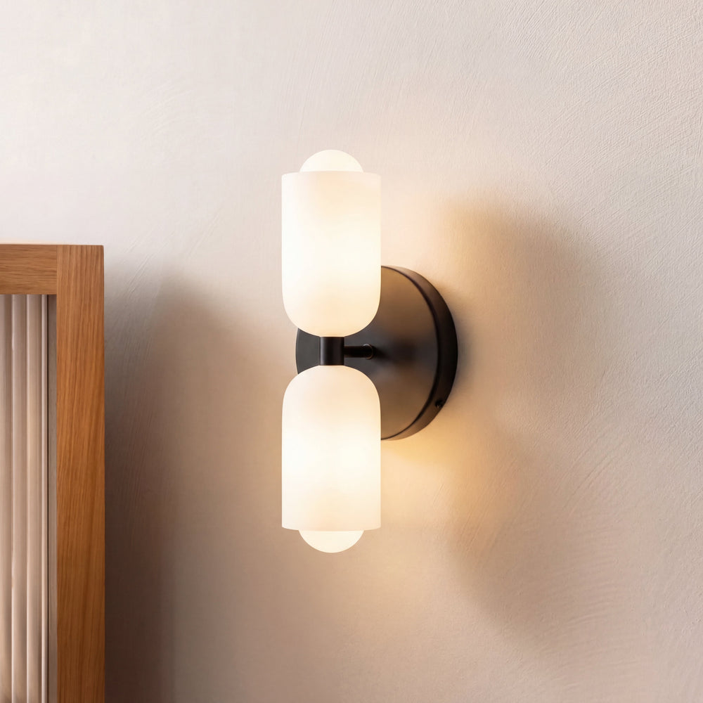 Side-angle view of the lit black wall light with two opal glass shades, mounted on a cream wall next to an oak frame. 2