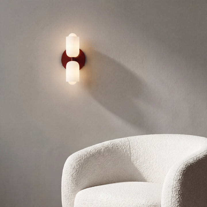 Lit wall light with a deep red backplate and white glass shades, casting a soft shadow on a gray wall above a white boucle armchair. 6