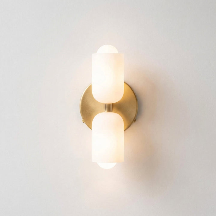 Front view of the wall light with a brushed brass backplate and two marbled white alabaster-style shades, glowing warmly on a white wall. 11