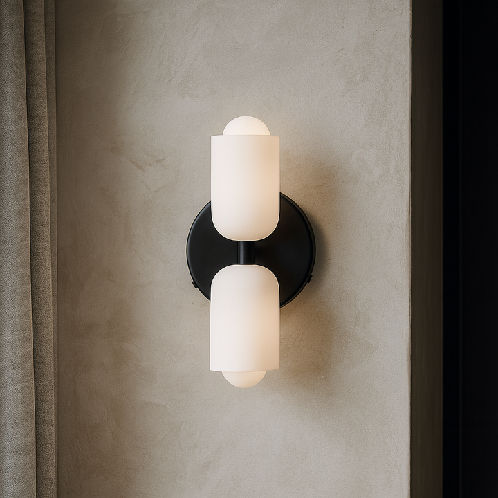 Black-backplate wall light with two frosted white glass shades, one pointing up and one down, glowing against a textured plaster wall beside a curtain. 1