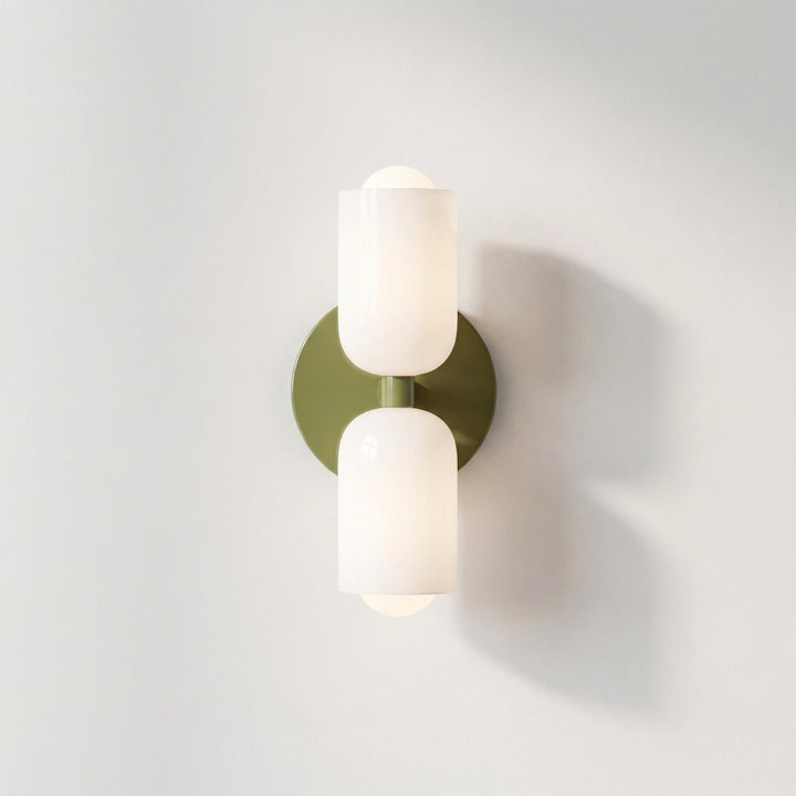 Front view of the wall light with an olive green backplate and two glossy white glass shades, casting a soft shadow on a white wall. 12