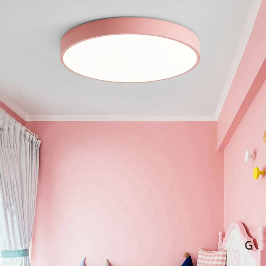 Pink round flush-mount ceiling light without wood trim, glowing in a pink children's bedroom with a curtain and decorative hooks.