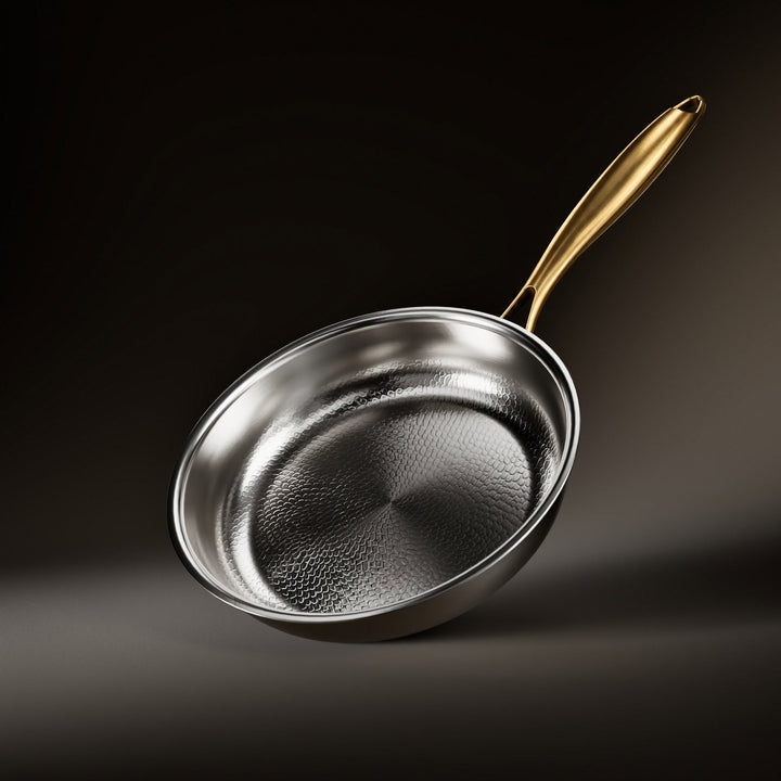 Tilted frying pan with a textured hammered silver cooking surface and a brushed gold-toned handle against a dark backdrop. 1