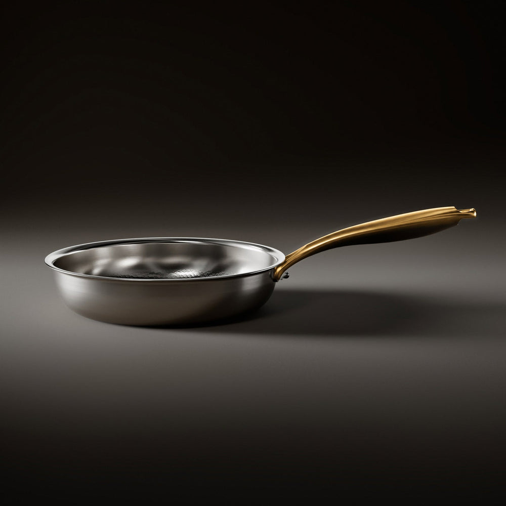 Side view of the silver frying pan resting on a dark surface, showing its low curved wall and gold-toned handle. 2