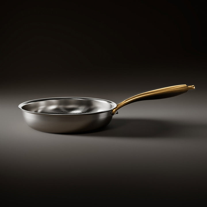 Side view of the silver frying pan resting on a dark surface, showing its low curved wall and gold-toned handle. 2