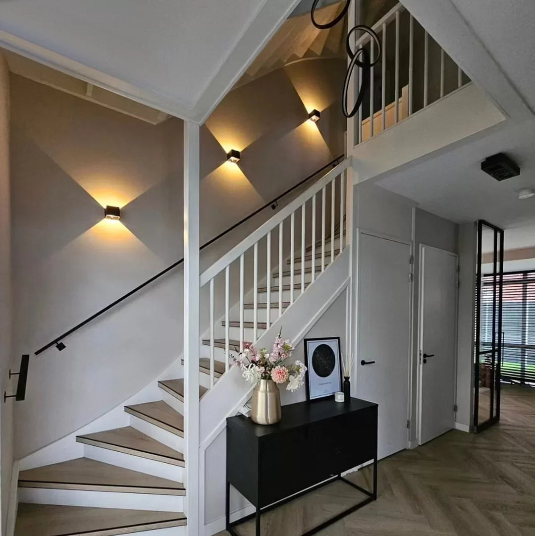 Three black cube wall lamps lighting a staircase wall in a modern hallway with white railings and a black console table. 7