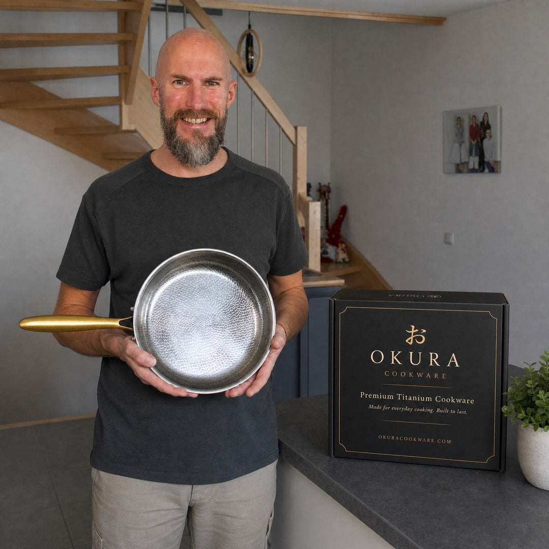 A smiling bald man with a beard holds one hammered pan in a home kitchen next to the black product box.