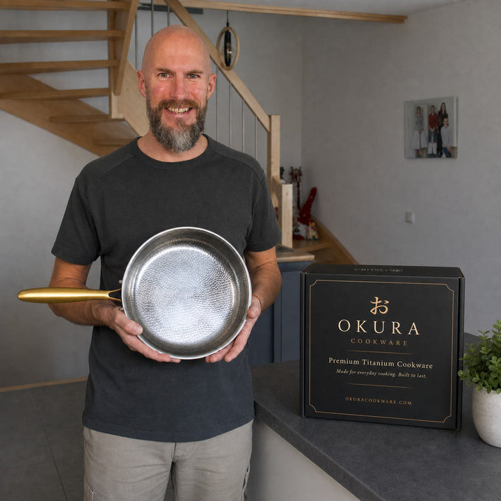 A smiling bald man with a beard holds one hammered pan in a home kitchen next to the black product box.