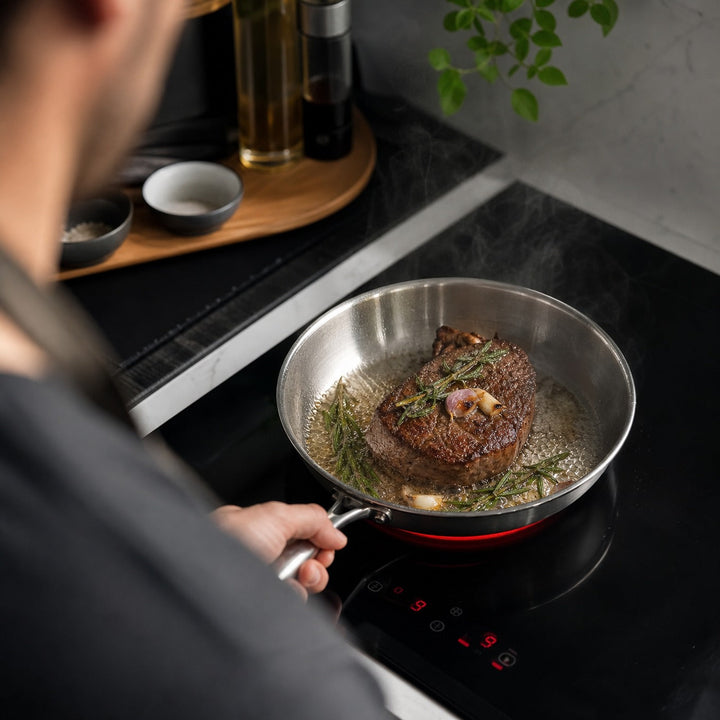 Man holding the pan by its handle over an induction cooktop while a steak sears with rosemary and garlic.