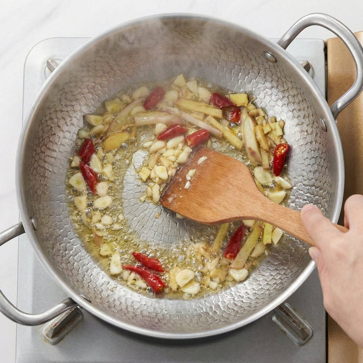 Detail of garlic, ginger and dried red chilies cooking in the stockpot with a wooden spatula.