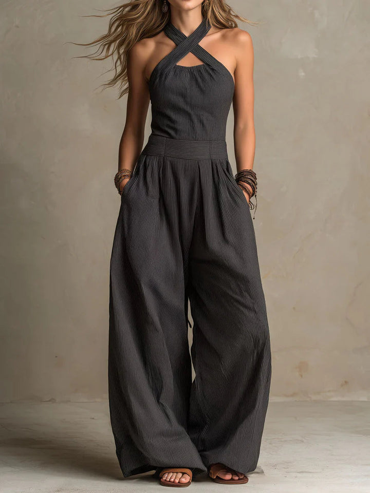 Model wears the pinstriped charcoal halter jumpsuit with crossed straps, hands in pockets, wide legs and sandals 1