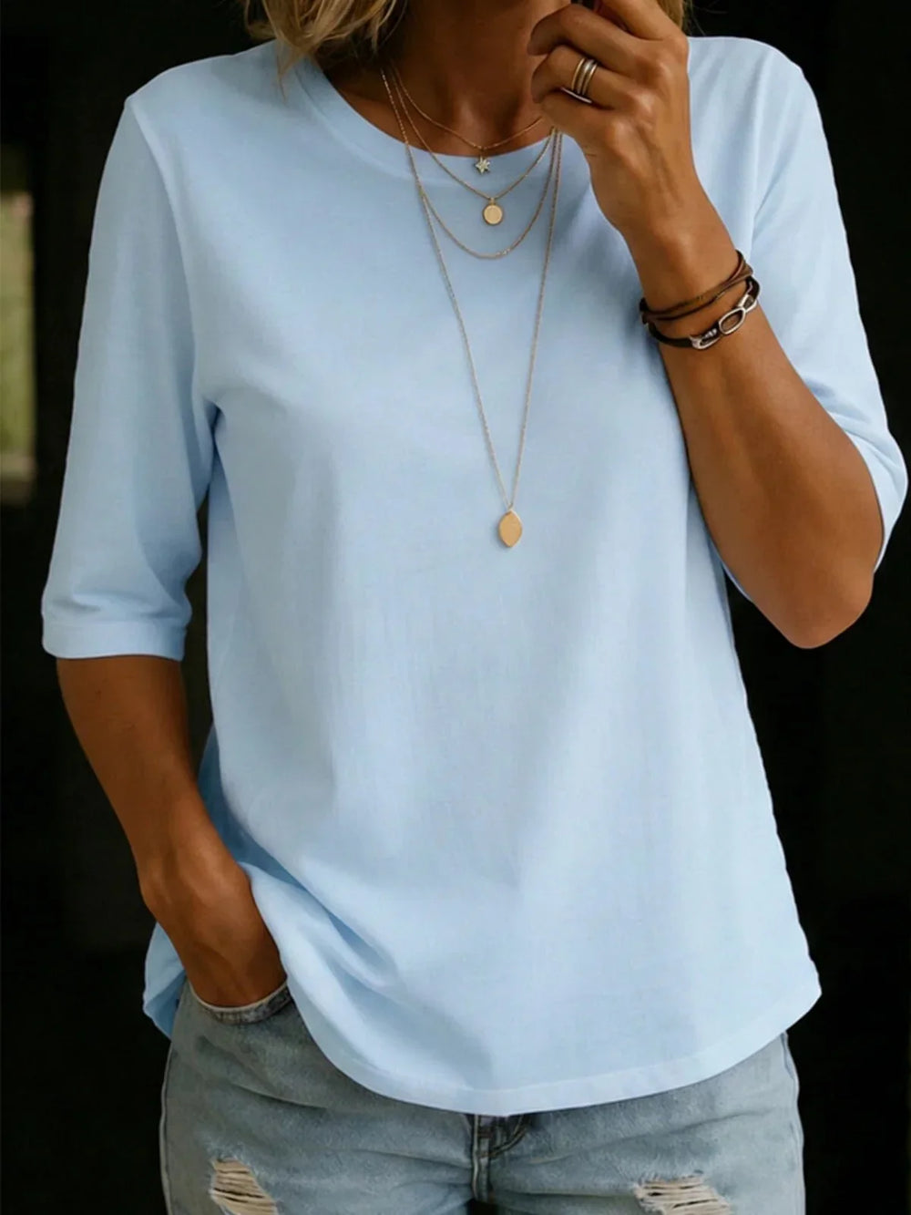 Woman wearing a light blue half-sleeve crew-neck tee with layered gold necklaces, bracelets and ripped light-wash jeans. 2