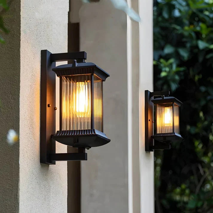 Two matching black lantern wall lights glowing warmly on pillars, with dark green foliage in the background. 2