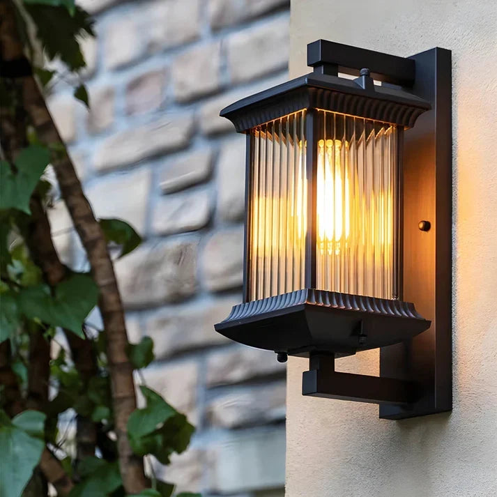 Close-up of the lit black lantern wall light mounted on a cream wall beside a stone wall and ivy leaves. 1