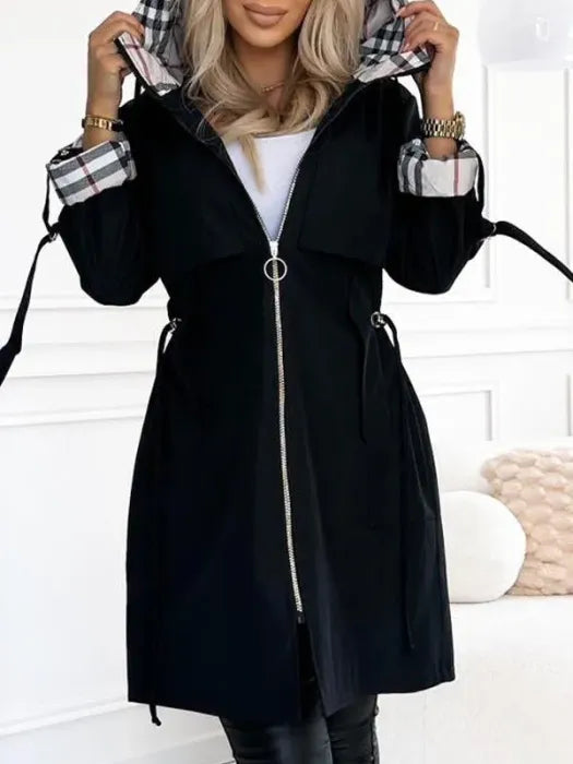 Woman wearing the black hooded jacket zipped up the front with a ring pull, hood raised to show plaid lining and cuffs.