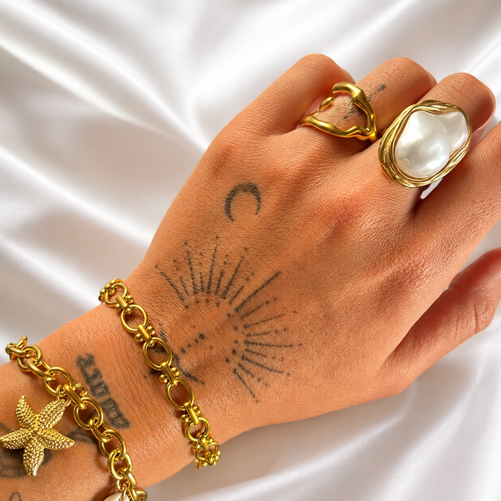 Tattooed hand on white satin wearing the wavy gold open ring beside a large pearl-style ring, with gold chain bracelets on the wrist. 4