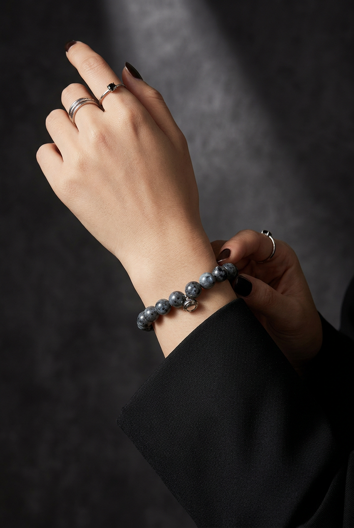 Woman's wrist wearing the gray speckled bead bracelet with silver rings and a black sleeve against a dark backdrop 4