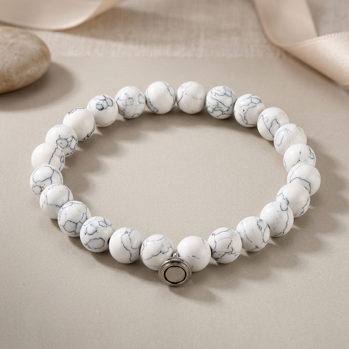 White bead bracelet with gray marbled veining and a silver clasp, set beside a pebble and ribbon 3