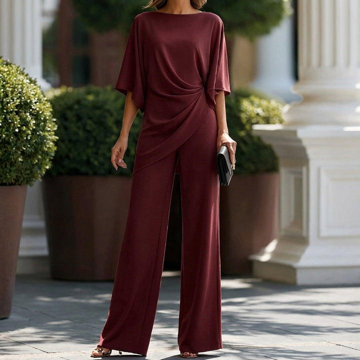 Model in the burgundy version, cropped at the face, showing the draped blouse and long wide-leg pants beside potted shrubs. 9