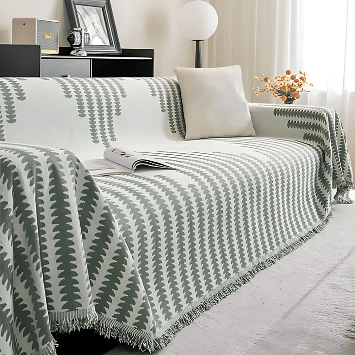 Sage green and cream graphic-print knit sofa cover another view, with a cushion, an open magazine and fringed trim.