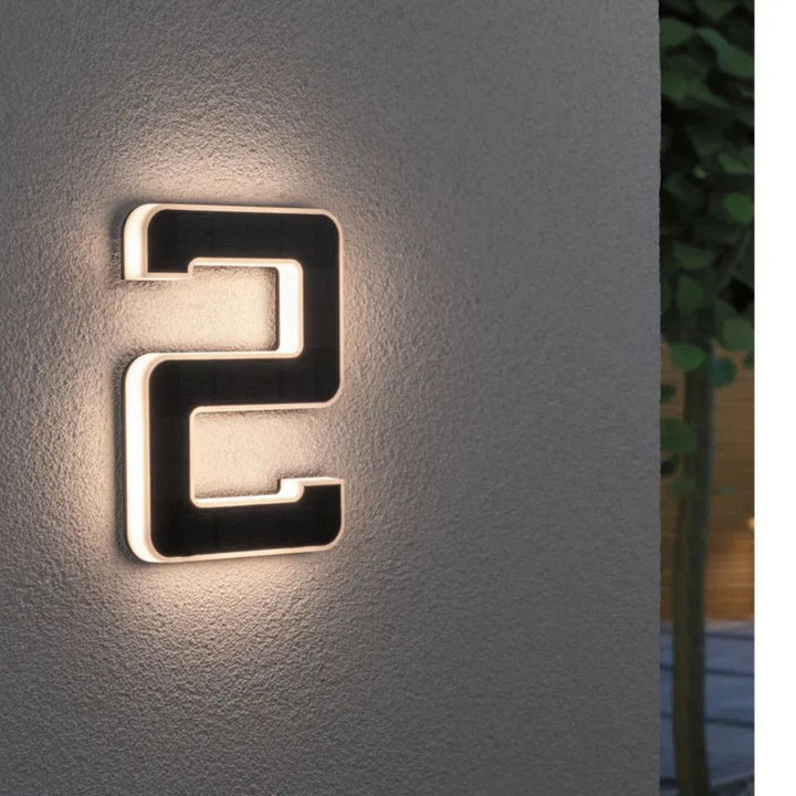 Solar illuminated house number 2 with warm LED outline and a dark solar panel face on a rough gray wall.