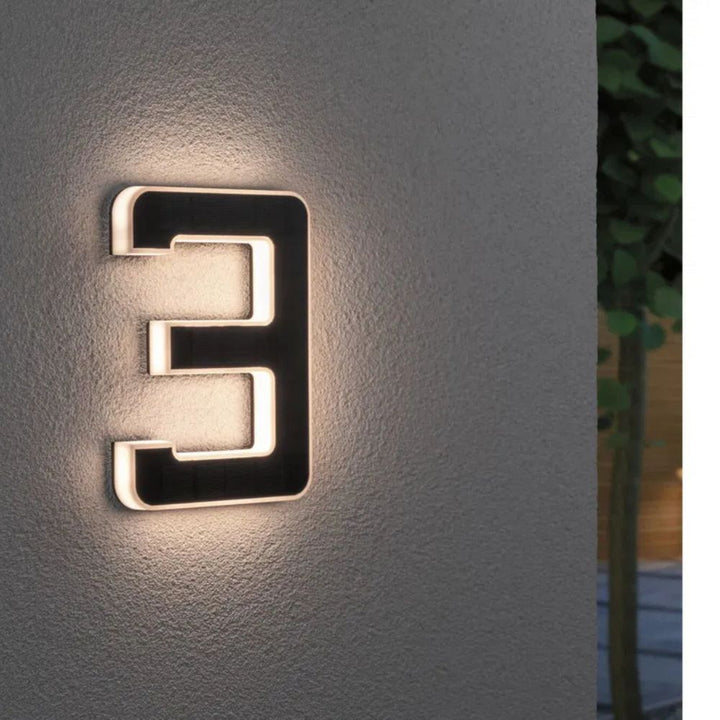Solar illuminated house number 3 with warm LED edge and a dark solar panel face on a gray wall.