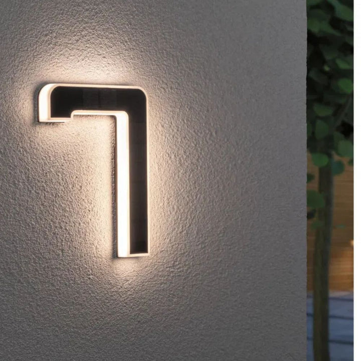 Solar illuminated house number 7 with warm LED edge light and a dark solar panel surface on a textured gray wall.