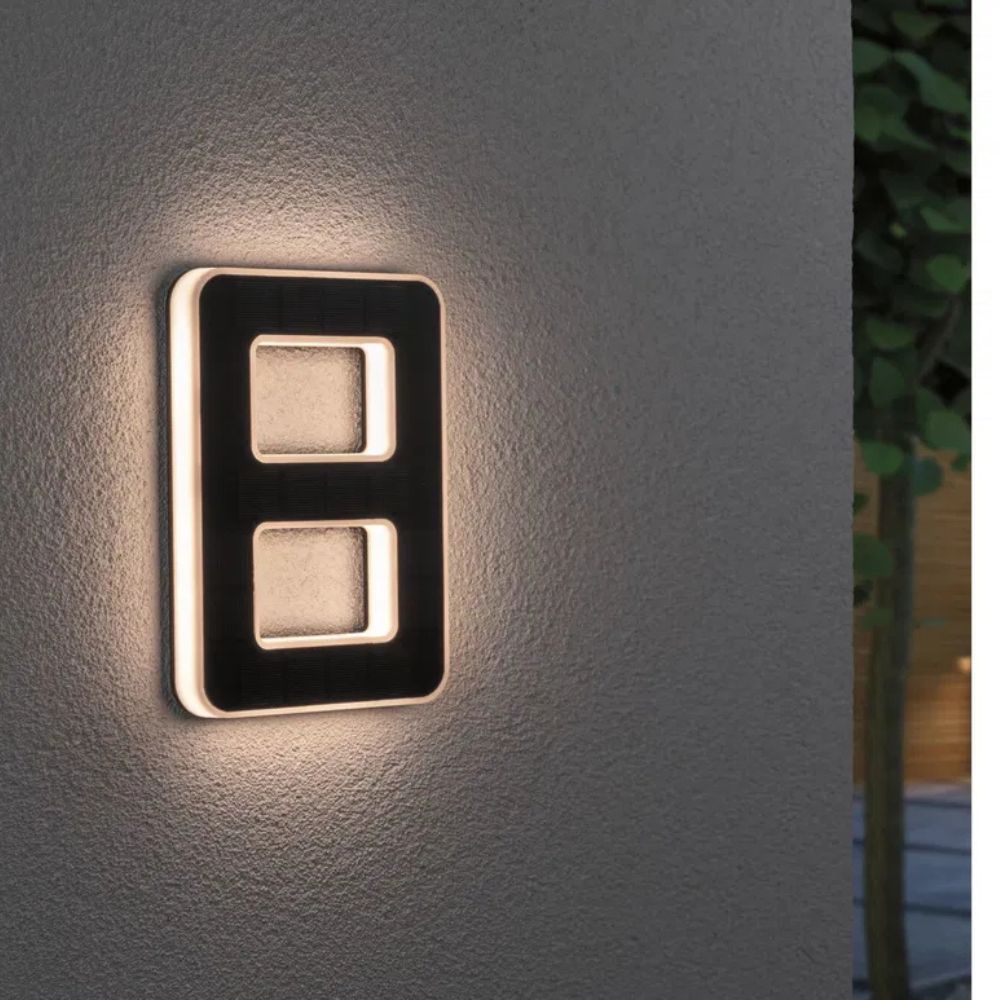 Solar illuminated house number 8 with warm LED edge and a dark solar panel face on a gray wall.