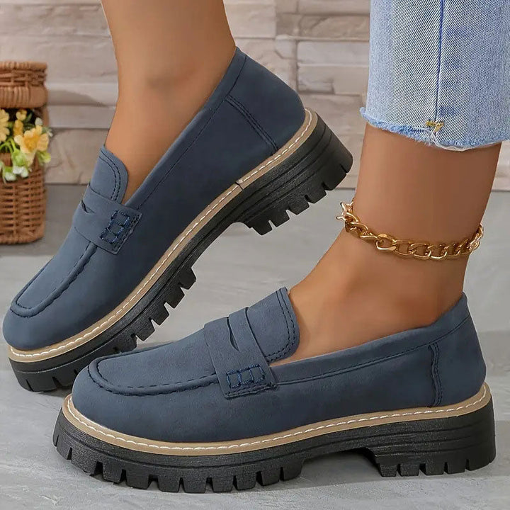 Navy blue loafers with penny straps and thick black platform soles worn with a gold chain anklet.