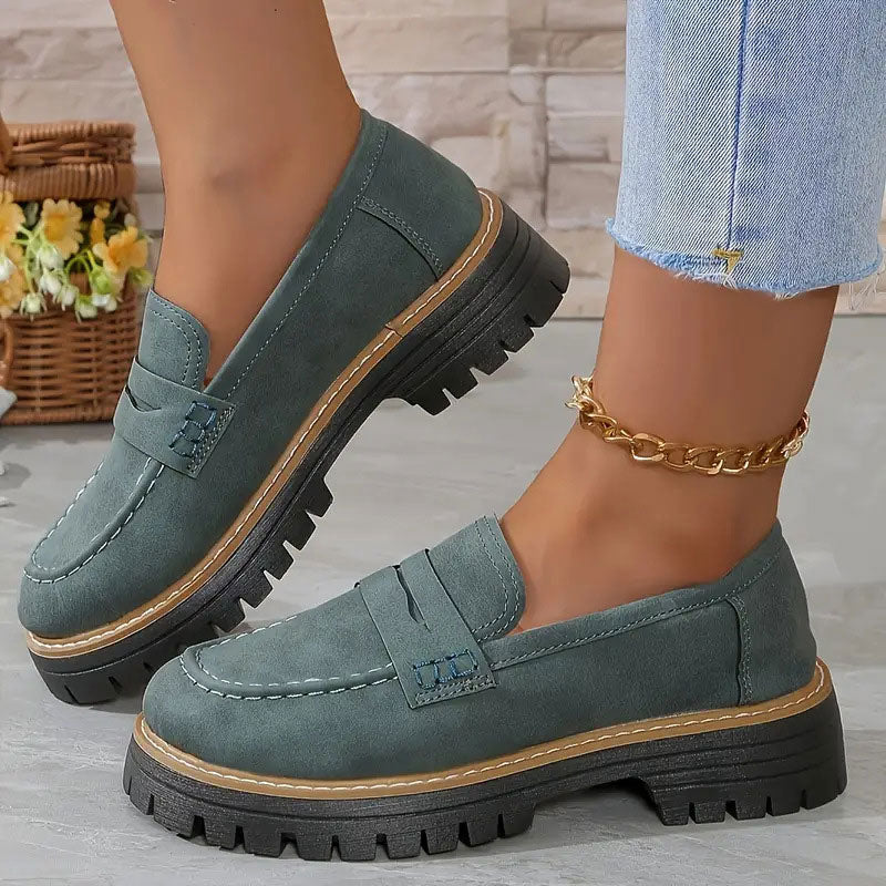 Muted green-gray loafers with tan welt and chunky black soles worn with a gold chain anklet.