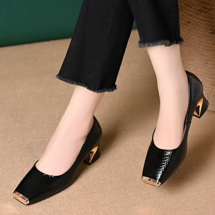 Model's crossed feet in black snake-embossed pumps with gold-tone square toe caps and sculpted heels, worn with frayed black cropped pants. 1