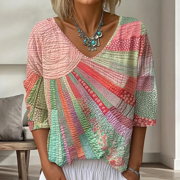 Close-up of the V-neck patchwork blouse in a soft pink, coral and mint green sunburst pattern, paired with white pants. 3
