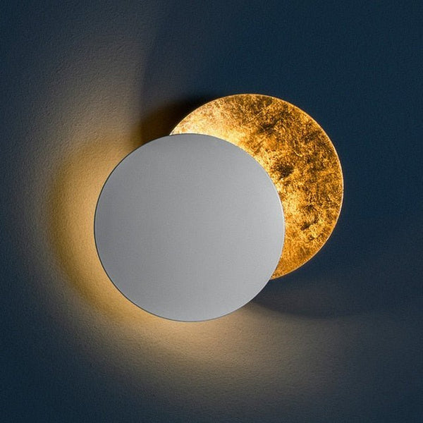 White front disc lamp with a gold-leaf backplate glowing warmly on a dark blue wall 8