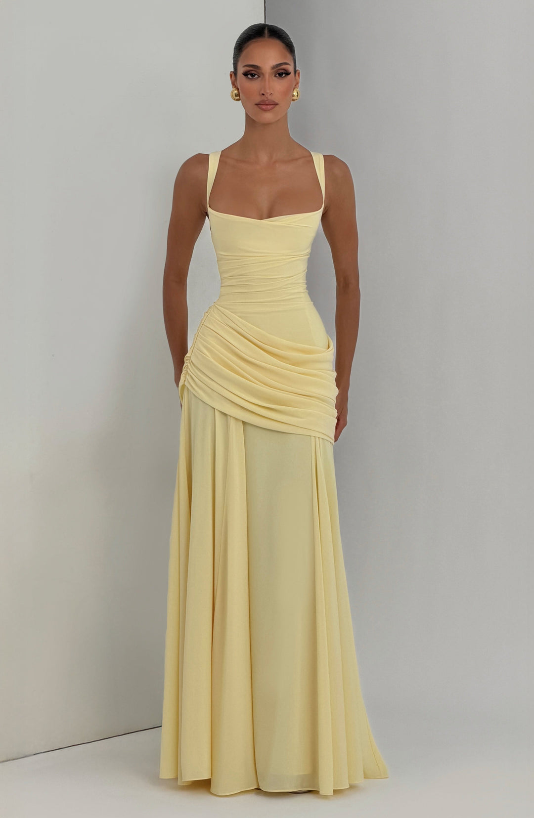 Pale yellow maxi dress worn against a gray corner backdrop, front view with ruched bodice and asymmetrical draped hip.