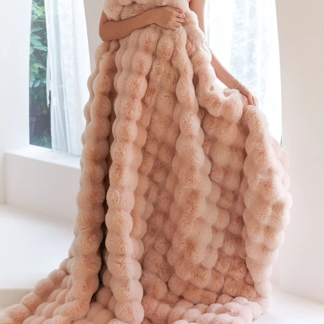 Woman holding a long peach-pink bubble-textured plush blanket that drapes to the floor beside a bright window 4