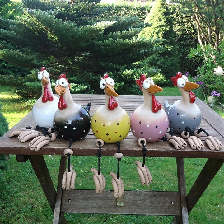 Five glazed ceramic chicken figurines in white, black, yellow, pink and gray with polka dots sit in a row on a wooden garden table. 1