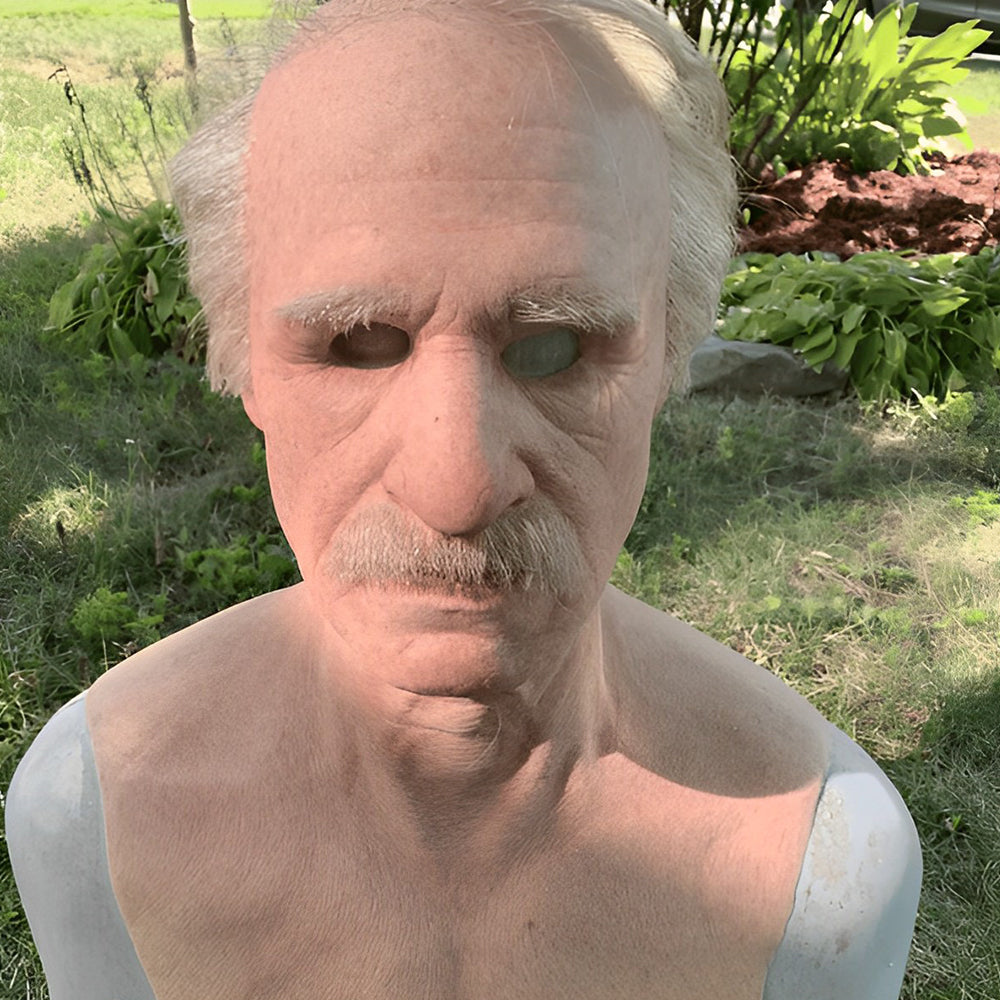 Mask of an older man with white hair and mustache on a chest form on a grassy lawn another view