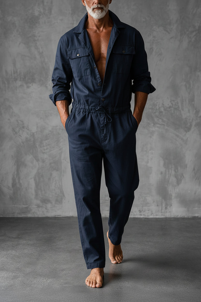 Barefoot man in a navy blue version of the jumpsuit, hands in pockets, gray backdrop 2
