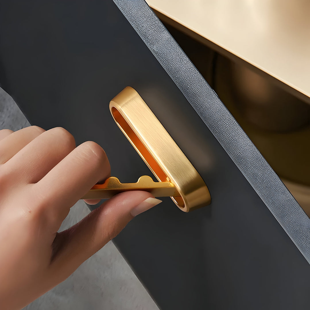 A hand pulling the folding arm out of a gold rounded Hidden Wall Hook mounted on a dark cabinet door.